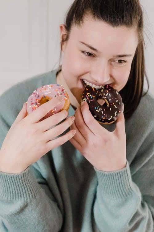 A Girl Biting into A Chocolate Glazed Donut While Holding a Strawberry Glazed Donut In other Hand