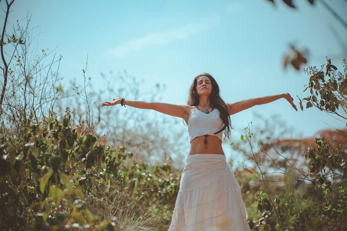Woman Wearing a White Top and Skirt Standing In A Wild Field with Both Arms Spread Out