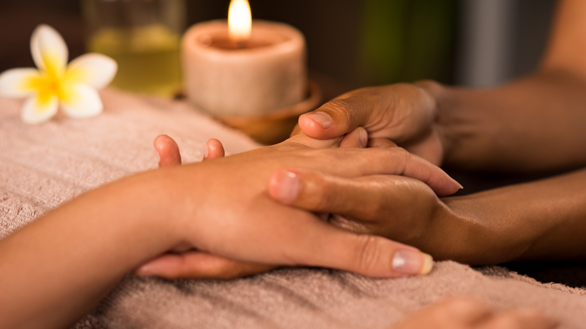 Close-up of two hands intertwined during a candlelit massage, with a warm massage oil candle and plumeria flower in the background.