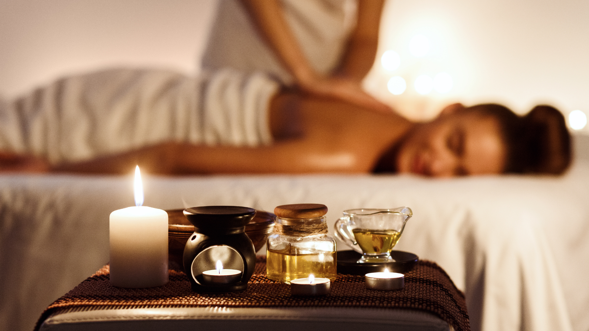 Lit massage candle and bottle of massage oil on a spa table with a relaxing massage in progress in the background